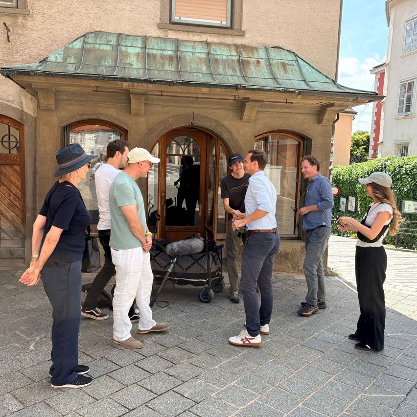 Eine Gruppe von Menschen steht vor einem Gebäude mit einem grünen Dach, einige unterhalten sich und andere schauen etwas an. Ein Mikrofon steht auf einem Trolley.