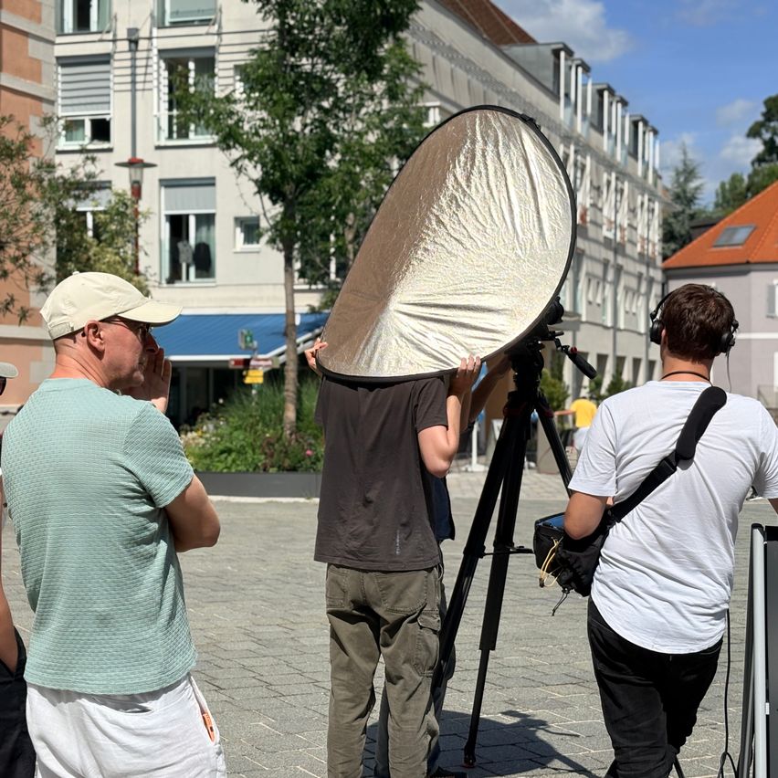 Drei Männer stehen auf einem gepflasterten Bürgersteig. Ein Mann hält einen großen kreisförmigen Lichtmodifikator über seinem Kopf. Ein anderer Mann trägt Kopfhörer und eine Kameratasche.