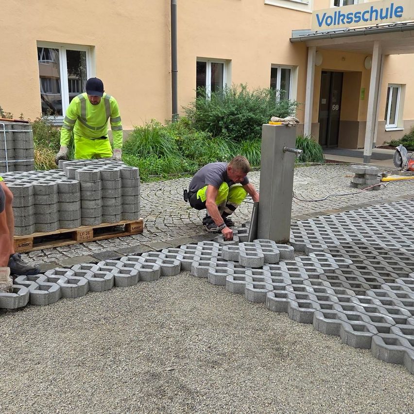 Zwei Arbeiter installieren graue sechseckige Betonblöcke auf einer Kiesoberfläche vor einem Gebäude mit einem Schild, das 'Volksschule' steht.