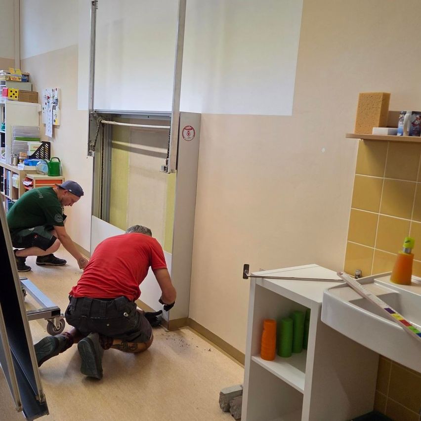 Zwei Männer arbeiten an einer Wand, einer kniet und hält ein Werkzeug, der andere steht und schaut auf die Wand. Dahinter befindet sich ein Regal mit verschiedenen Gegenständen.