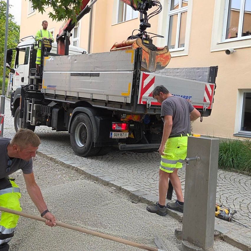 Zwei Männer in Warnwesten arbeiten auf der Straße, einer benutzt einen Hammer und der andere steht neben einem LKW mit einem Kran. Ein Gebäude mit Fenstern und einer Überwachungskamera ist im Hintergrund.