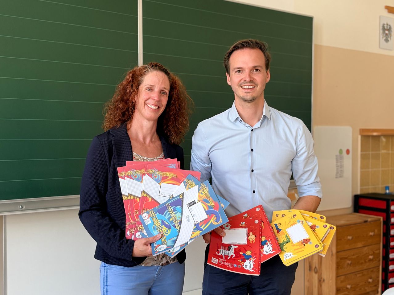 Ein Mann und eine Frau stehen vor einer grünen Tafel, halten bunte Notizbücher in den Händen und lächeln für die Kamera.