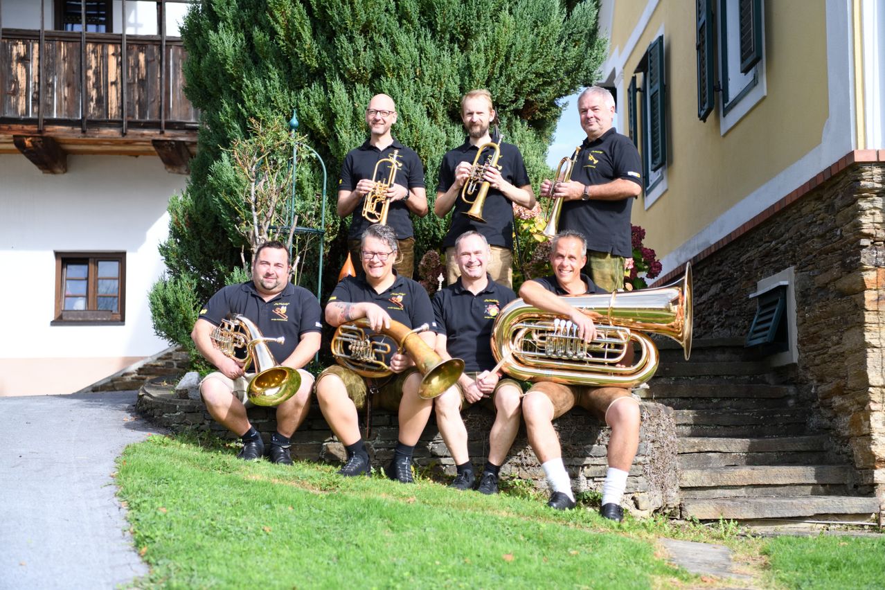Eine Gruppe von Männern in schwarzen Shirts und Shorts posiert mit ihren Blechblasinstrumenten in einem Garten für ein Foto.