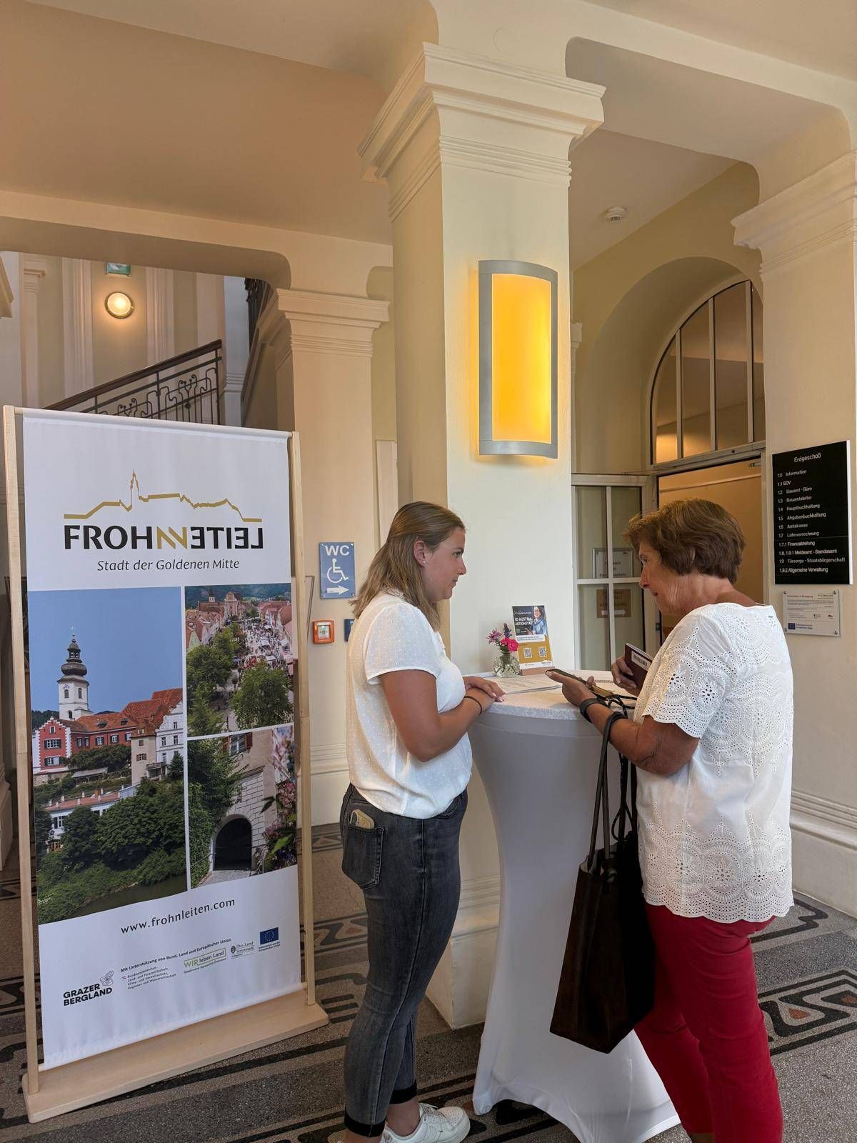 Zwei Frauen stehen an einer Rezeption in einer Lobby, eine schaut in ein Buch und die andere hält ein Buch. Ein Banner mit dem Text 'FROHNLEITER' und einem Bild einer Stadt steht hinter ihnen.