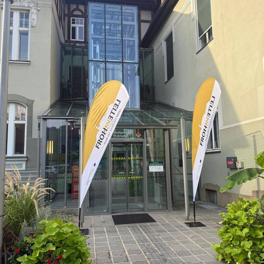 Der Eingang zu einem modernen Gebäude mit Glaswänden und Türen, zwei gelben Bannern mit weißem Text. Das Gebäude hat einen Balkon und mehrere Fenster. Im Vordergrund befinden sich Pflanzen und Grünflächen.