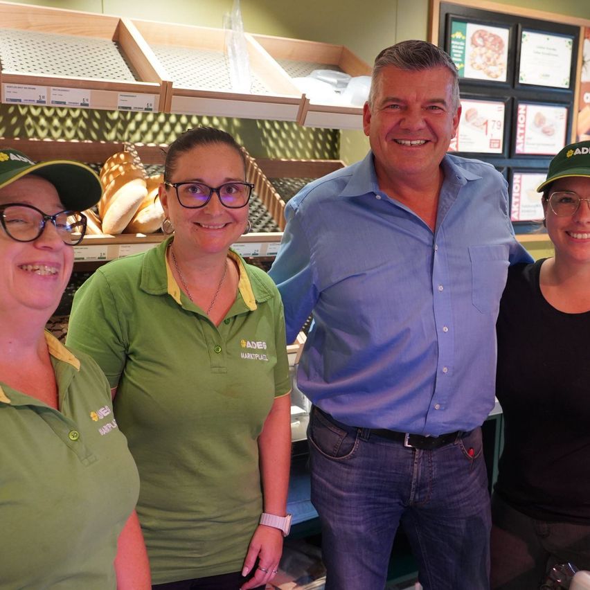 Drei Personen stehen vor einer Bäckereitheke und lächeln. Ein Mann in einem blauen Hemd und zwei Frauen in grünen Hemden. Dahinter befinden sich Regale mit Brot und eine Menütafel.