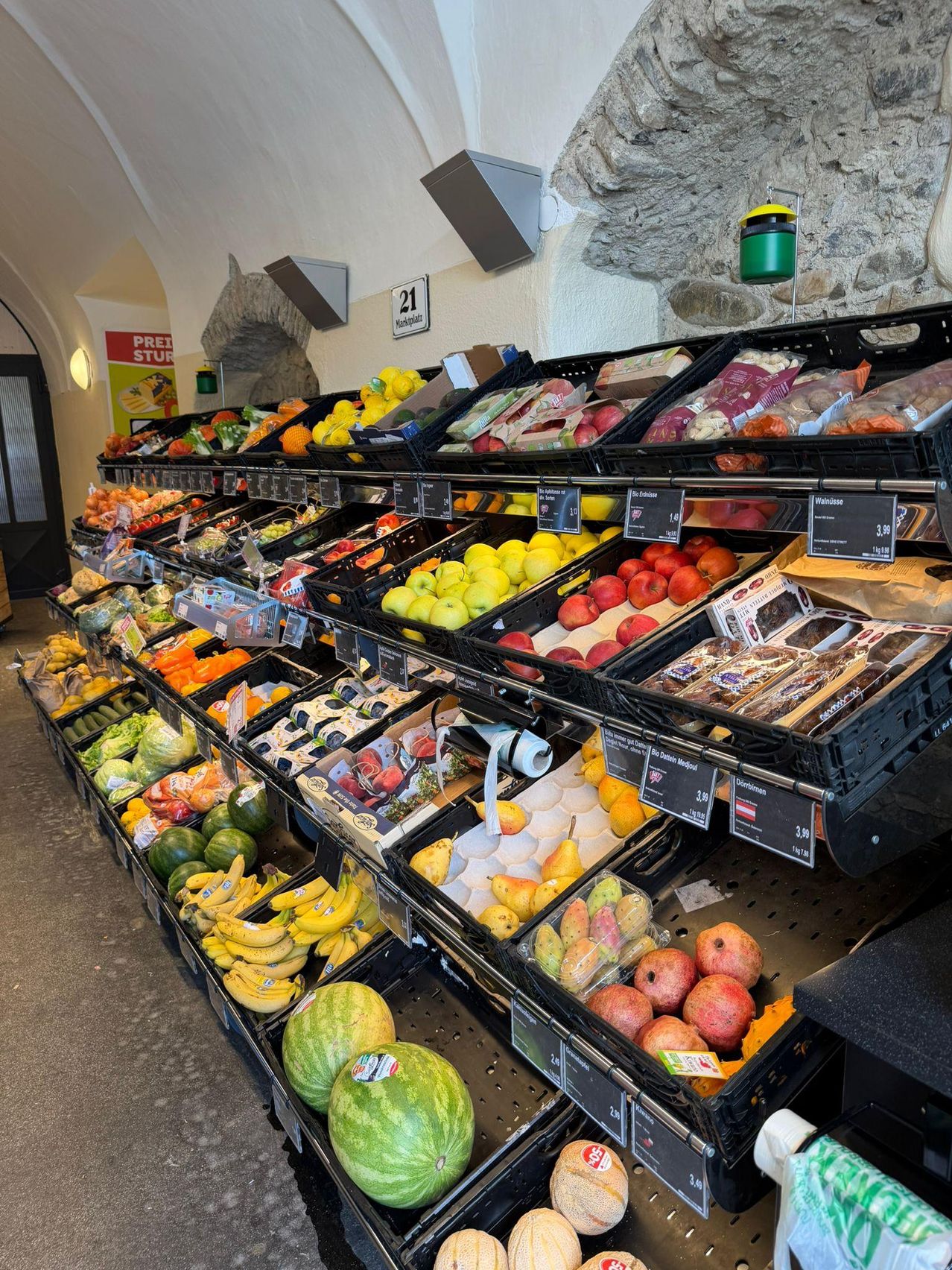 Ein Lebensmittelregal mit verschiedenen Obst- und Gemüsesorten, darunter Äpfel, Bananen, Wassermelonen und Birnen, alles ordentlich in schwarzen Kisten mit Preisschildern angeordnet.