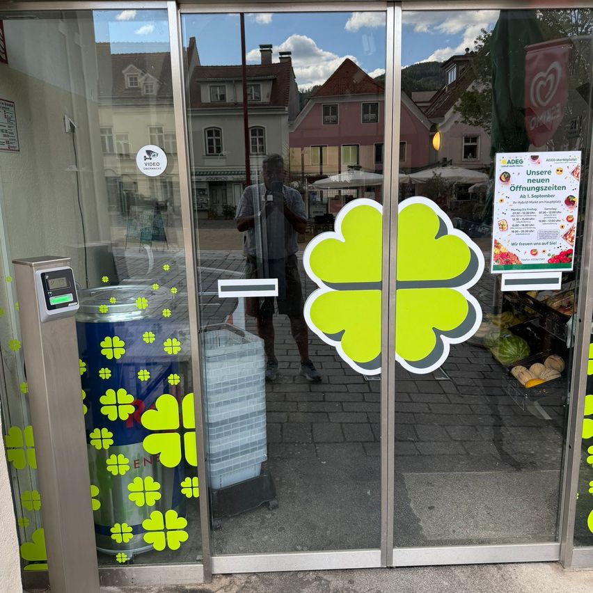 Eine Glastür mit einem grünen vierblättrigen Kleeblatt-Design, einem Videoüberwachungsaufkleber und einem Plakat, das neue Eröffnungen ankündigt. Ein Mann ist in der Scheibe reflektiert.