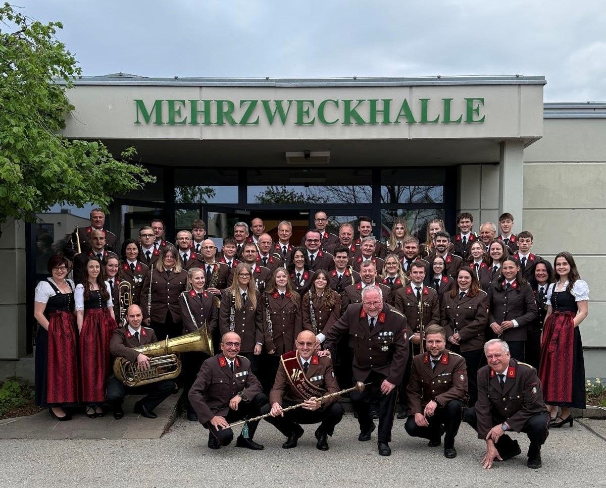 Eine große Gruppe von Menschen in Uniform, einschließlich Männer und Frauen, posiert für ein Foto vor einem Gebäude mit dem Schild 'Mehrzweckhalle'. Einige halten Musikinstrumente.