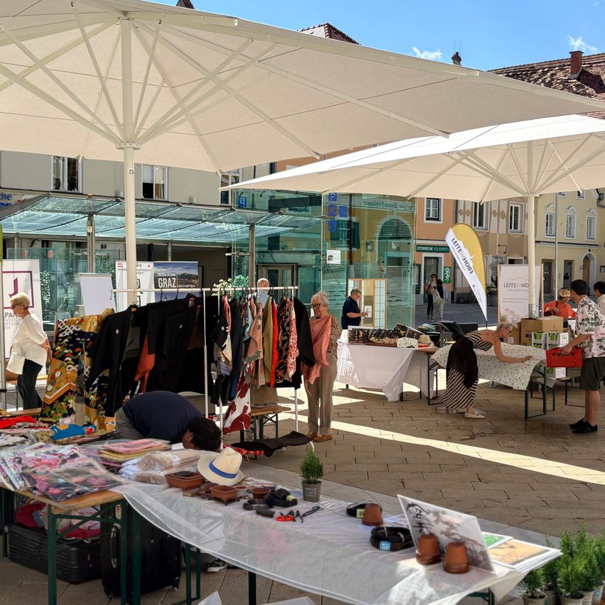 An outdoor market with various stalls under large umbrellas. People are browsing through clothes and other items. The market is set up in a sunny area with buildings in the background.