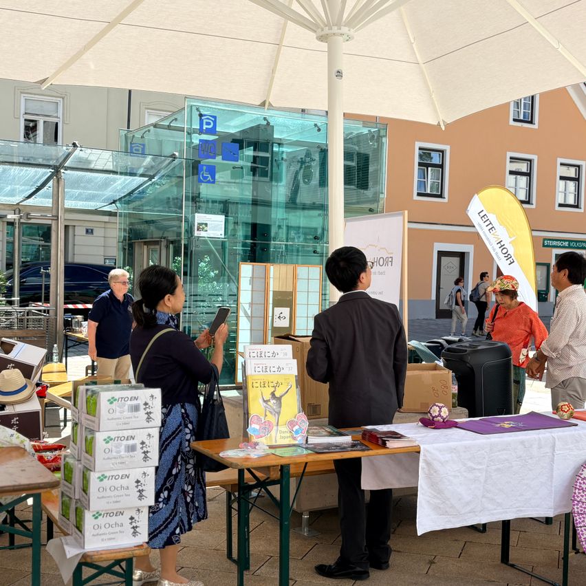 A group of people are gathered under an umbrella at a market, with one woman holding a phone and another man looking at a table with various items.