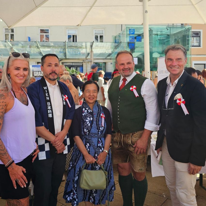 Five individuals stand under an umbrella, smiling for a photo. The man on the right wears a green vest and brown shorts. The woman in the middle holds a green handbag. Behind them, buildings and people are visible.