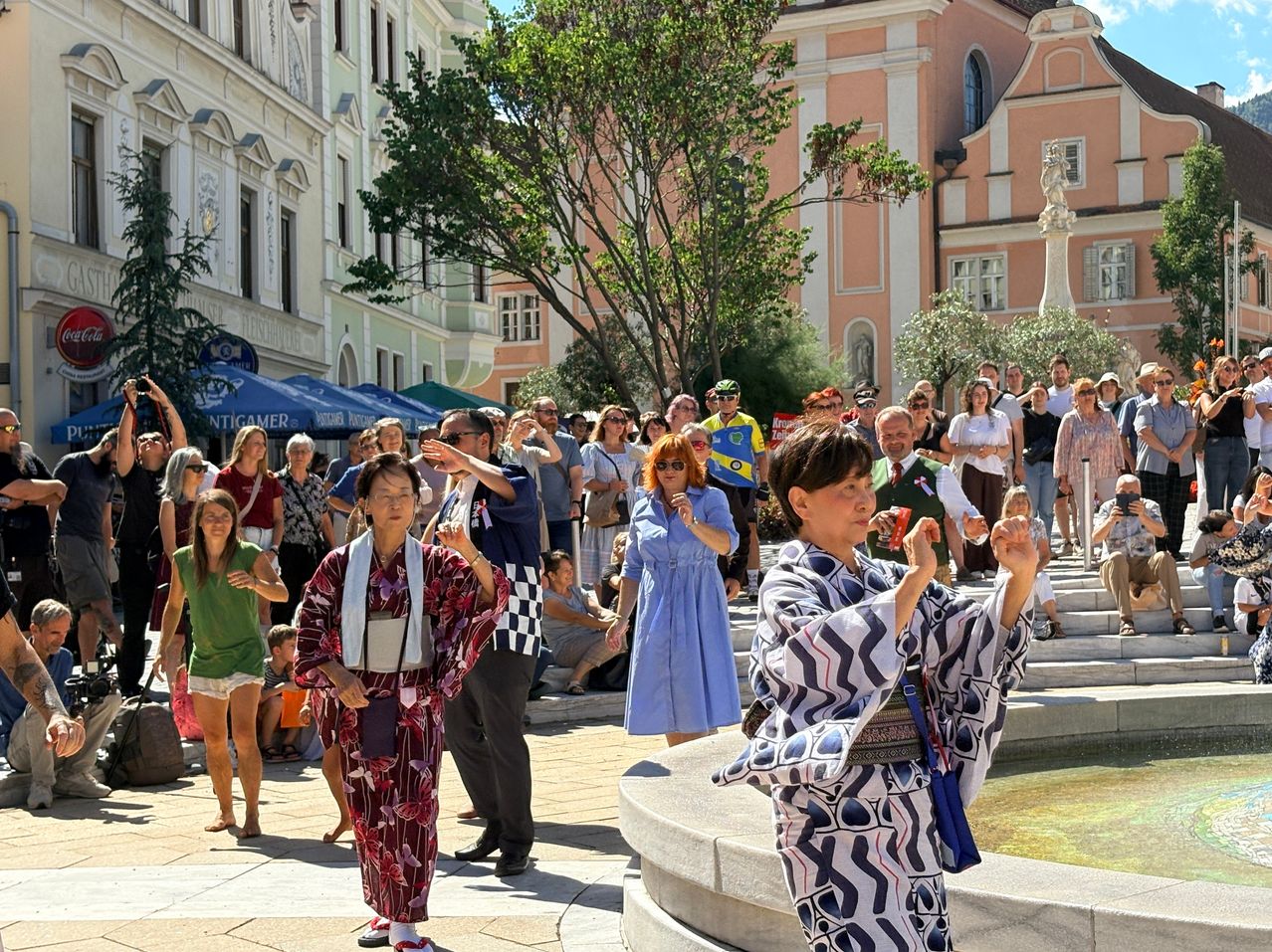 A group of people, some in traditional Japanese attire, are gathered in an outdoor urban setting, possibly participating in a cultural event.