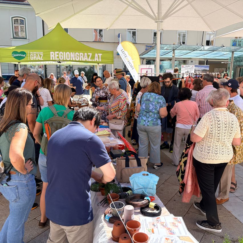 A crowd of people are gathered around a table with various items on display under a green tent.