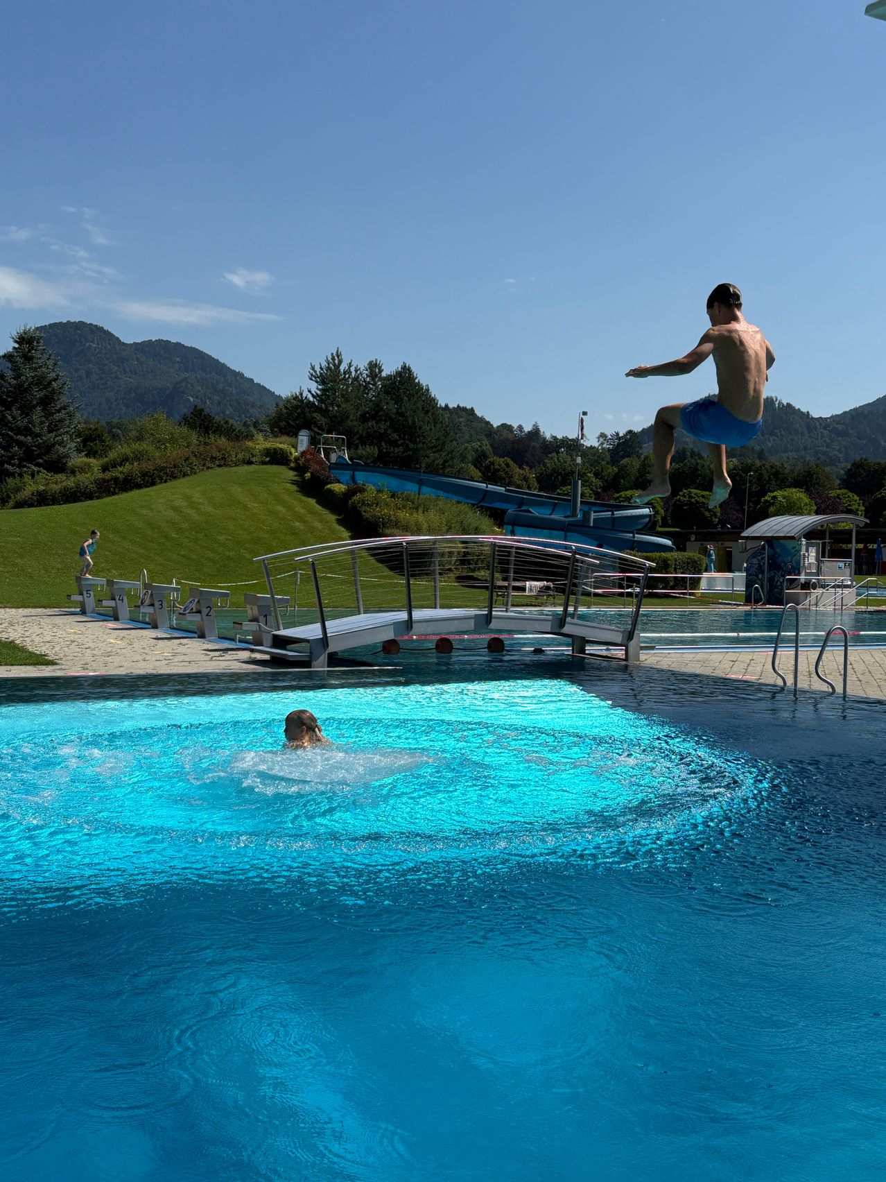 Ein Junge springt in einen Pool, während ein anderer darunter schwimmt. In der Nähe ist eine Wasserrutsche zu sehen. Im Hintergrund sind eine Wiese und Berge zu erkennen.