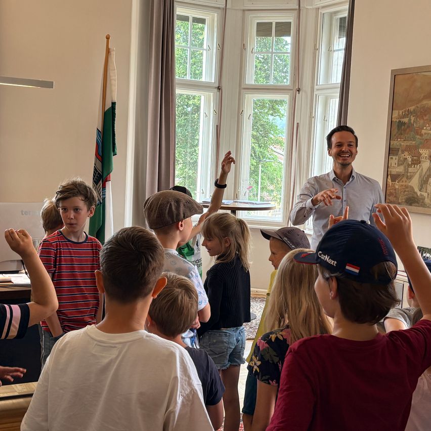 Ein Mann unterrichtet eine Gruppe von Kindern in einem Raum. Die Kinder tragen Freizeitkleidung und einige Mützen. Eine Fahne steht in der Ecke des Raumes.
