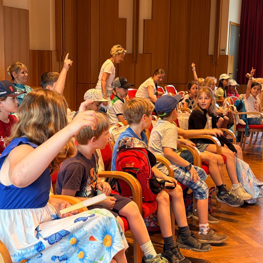 Ein Klassenzimmer mit Kindern und Erwachsenen, die sitzen, einige heben die Hände, eine Frau steht in der Nähe einer Tür und eine Person liest im Vordergrund.