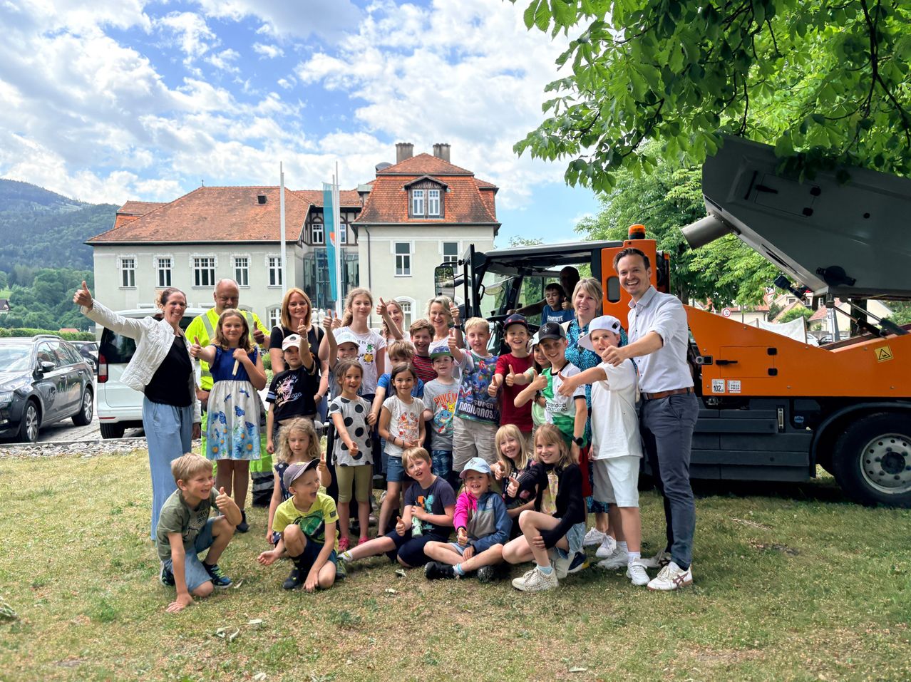Eine Gruppe von Kindern, Erwachsenen und einem Mann posiert für ein Foto vor einem Gebäude mit einem Baufahrzeug.