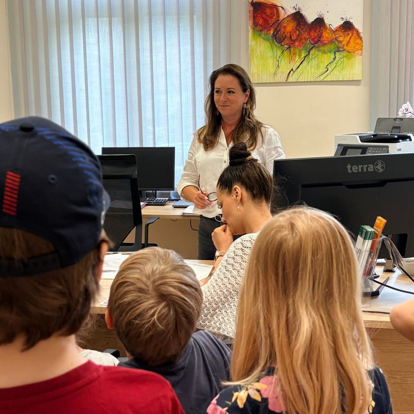 Eine Frau steht vor einem Schreibtisch mit Monitoren und Papieren und unterrichtet eine Klasse von Kindern, die auf Stühlen sitzen.