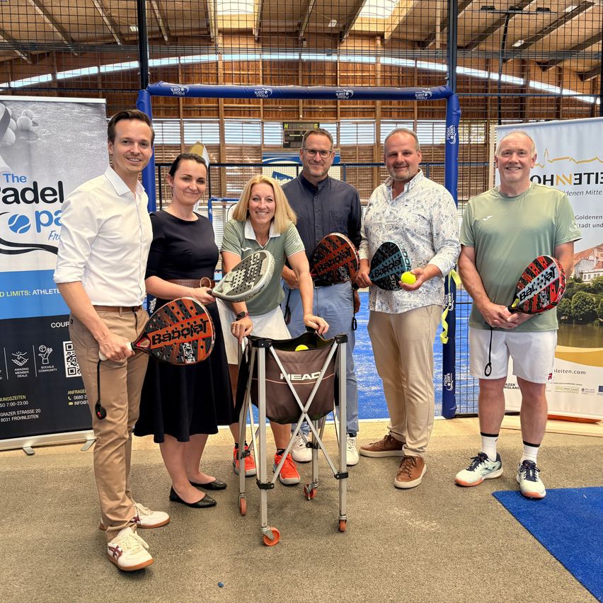 Fünf Menschen posieren für ein Foto auf einem Paddle-Tennisplatz. Zwei von ihnen halten Schläger. Der Mann auf der linken Seite trägt ein weißes Hemd, während der Mann auf der rechten Seite ein grünes Hemd trägt. Hinter ihnen befindet sich ein blauer Banner.