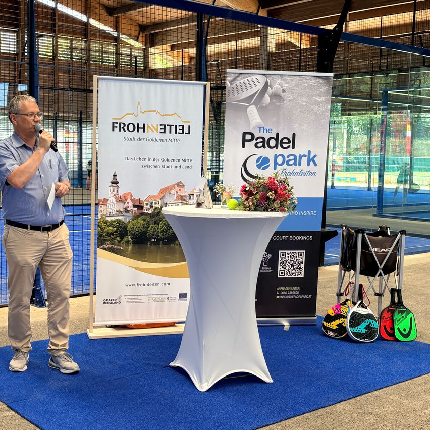 Ein Mann hält eine Rede in einer Sporthalle mit Tennisausrüstung auf einem Tisch und zwei Bannern. Die Banner lauten 'FROHNLEITEN' und 'The Padel park Erholen'.