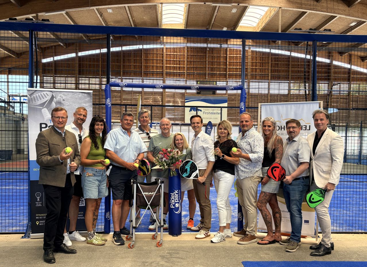Eine Gruppe von Menschen steht in einer Sporteinrichtung, hält Tennisschläger und Tennisbälle und posiert für ein Foto. Der Bereich ist von einem blauen Netz und einem Zaun umgeben.