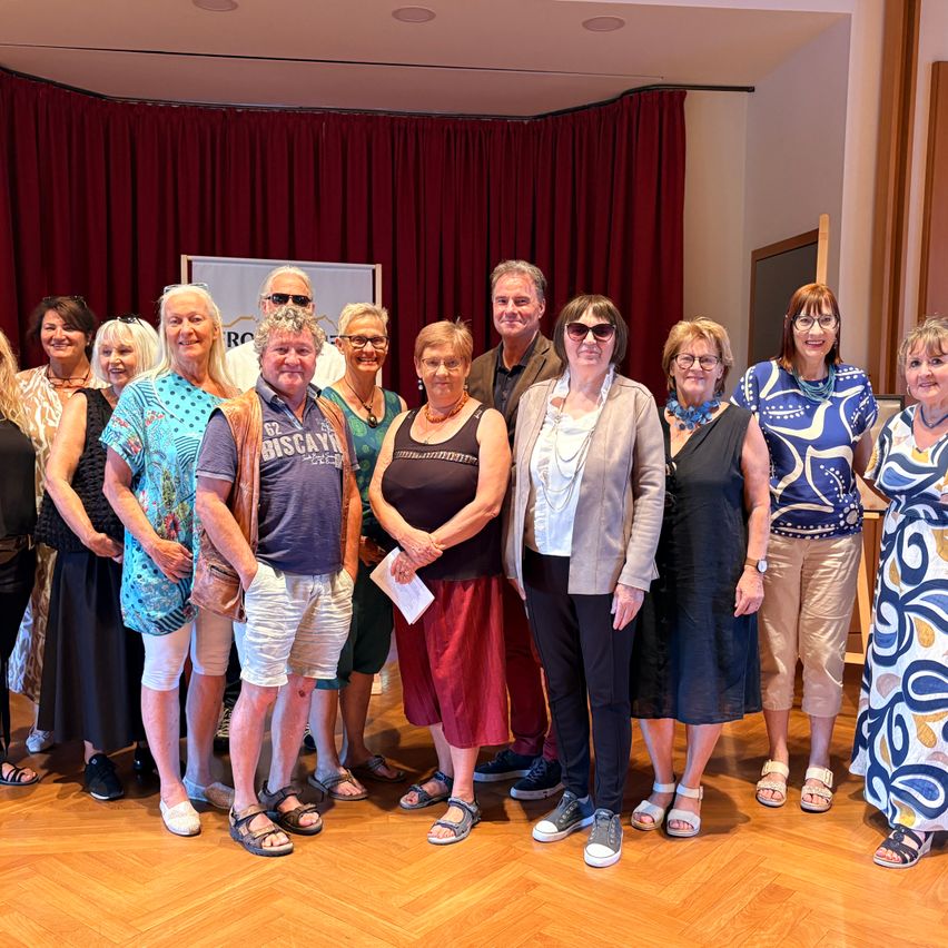 Eine Gruppe von Menschen, die vor einem roten Vorhang für ein Foto posieren. Sie tragen Freizeitkleidung und einige halten Papiere.