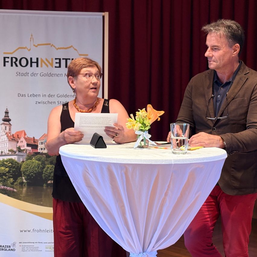 Ein Mann und eine Frau stehen hinter einem Tisch und schauen sich an. Die Frau hält ein Papier und einen kleinen Blumenstrauß. Hinter ihnen ist ein Banner mit einem goldenen Stadtbild.