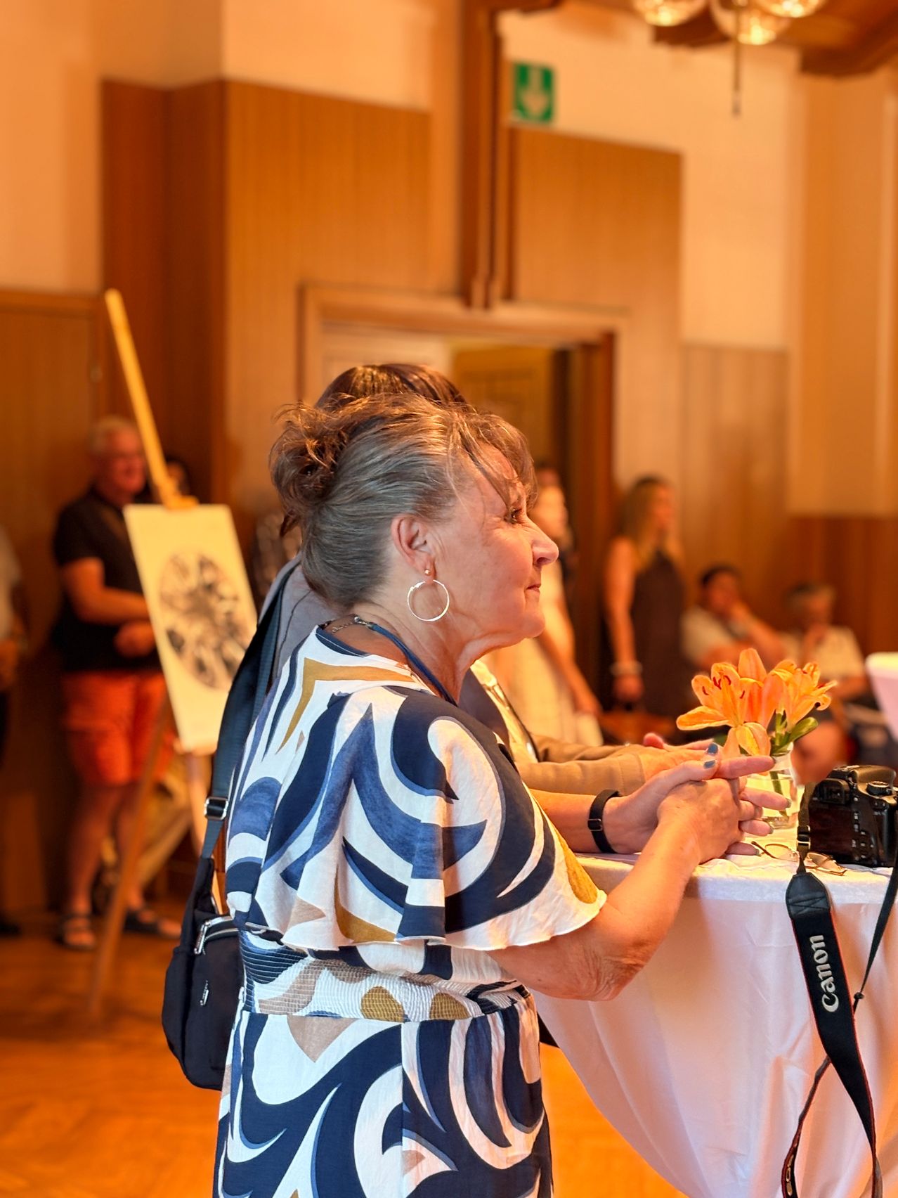 Eine ältere Frau mit Blumen steht vor einem Tisch mit einer Kamera, während ein Mann auf einer Leinwand zeichnet. Mehrere Menschen stehen herum, möglicherweise bei einer Veranstaltung.