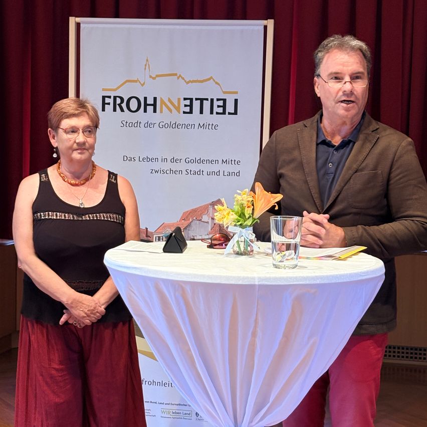 Eine ältere Frau und ein Mann stehen vor einem Tisch mit Blumen. Der Mann spricht und ein Banner mit den Worten Frohnleiten steht hinter ihnen.
