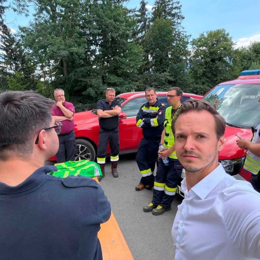 Mehrere Feuerwehrleute und andere Personen stehen um ein rotes Auto. Ein Mann spricht in ein Telefon.