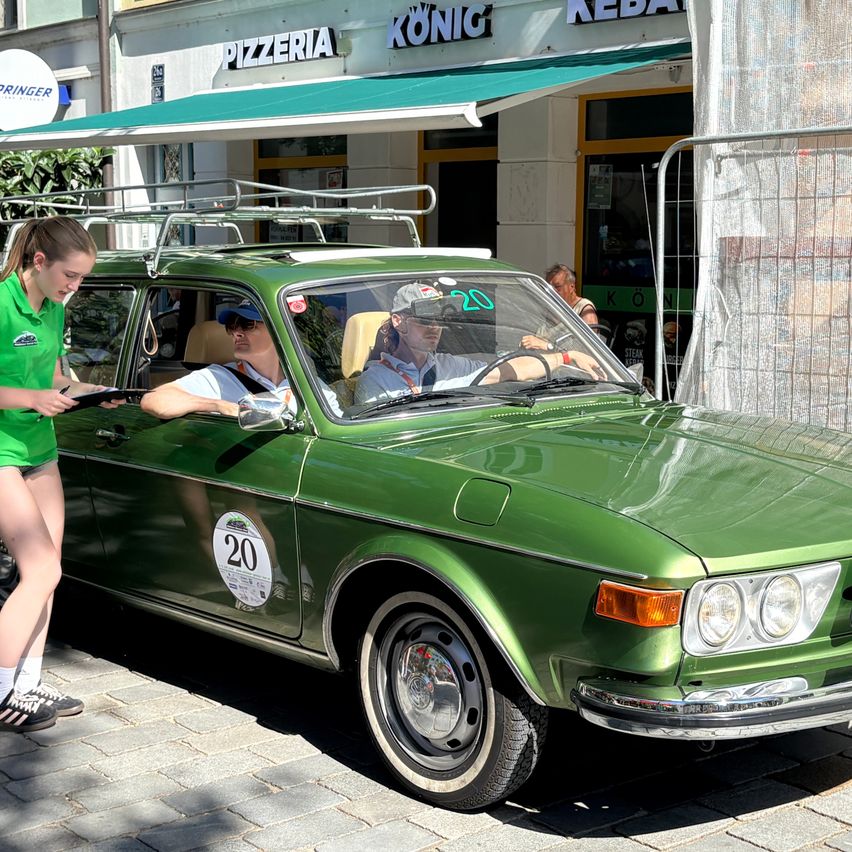 Ein grünes Oldtimer-Auto mit der Nummer 20 ist auf der Straße geparkt. Eine Frau in einem grünen Shirt steht neben dem Auto. Zwei Männer sitzen im Auto. Dahinter befindet sich ein Gebäude mit einem grünen Vordach.