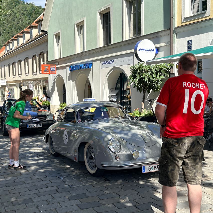 Ein silberner Vintage-Porsche ist in der Stadt geparkt. Ein Mann in einem Rooney 10-Shirt steht neben ihm. Eine Frau mit grünem Shirt ist in der Nähe. Gebäude und Bäume sind im Hintergrund.