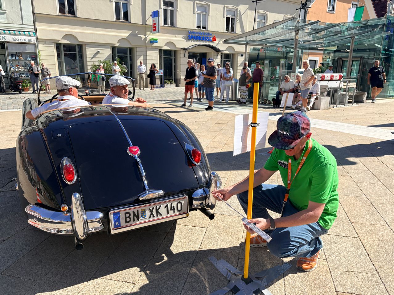 Ein Mann überprüft die Rückseite eines Oldtimers, während sich andere in der Nähe eines Gebäudes versammeln.