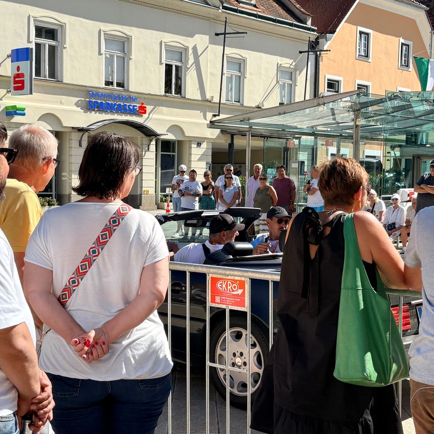 Eine Gruppe von Menschen hat sich um ein Cabriolet mit einem roten Schild davor versammelt, in der Nähe einer Metallbarriere. Sie scheinen etwas vor sich zu beobachten. Im Hintergrund befinden sich Gebäude.