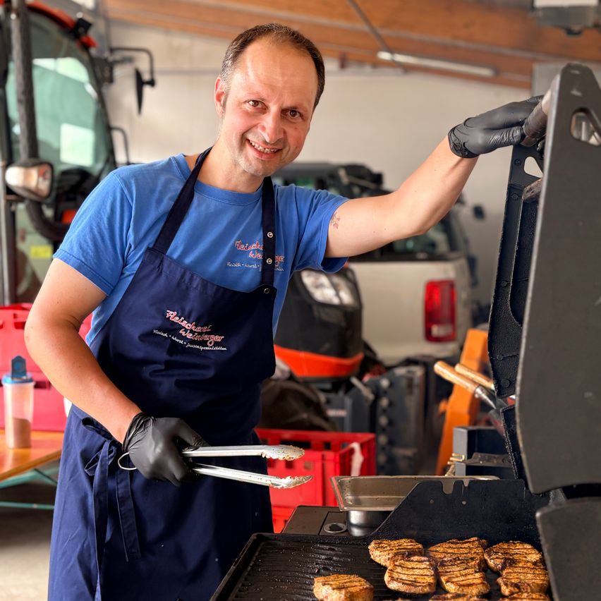 Ein Mann in einem blauen Schürze mit Text und schwarzen Handschuhen grillt Fleisch auf einem Grill in einer Garage mit Fahrzeugen im Hintergrund.