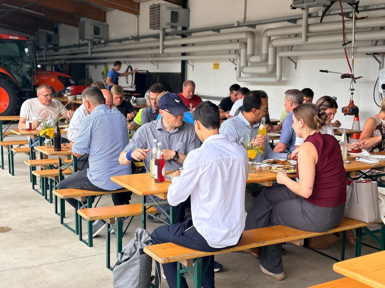Eine Gruppe von Menschen sitzt auf Holzbänken in einem großen Innenbereich, möglicherweise in einem Restaurant. Sie essen und trinken, in der Nähe befinden sich einige Tische und Stühle.
