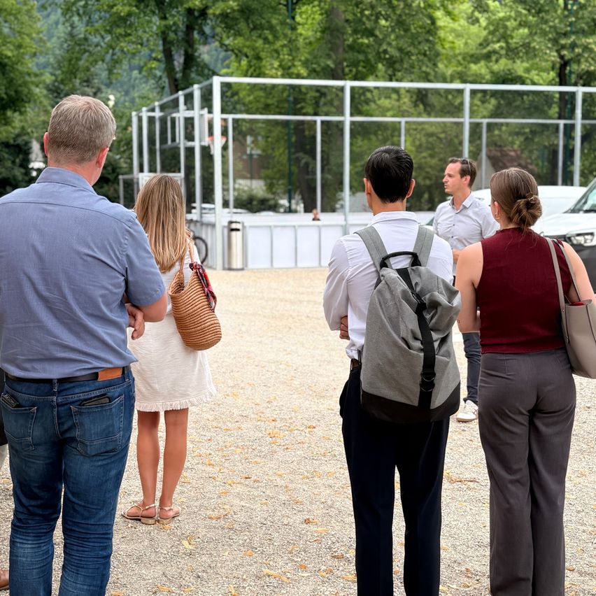 Fünf Personen, zwei Männer und drei Frauen, stehen draußen in der Nähe eines Metallzauns, wobei eine Frau einen Korb hält und ein Mann einen Rucksack trägt. Bäume und ein Fahrzeug sind im Hintergrund.
