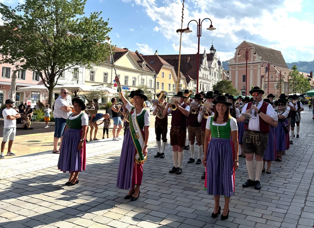 Eine Gruppe von Musikern in traditioneller bayerischer Kleidung steht auf einer Kopfsteinpflasterstraße und tritt auf. Sie halten Blechblasinstrumente und einige tragen Schärpen. Zuschauer stehen hinter ihnen. Gebäude mit Balkonen sind im Hintergrund.