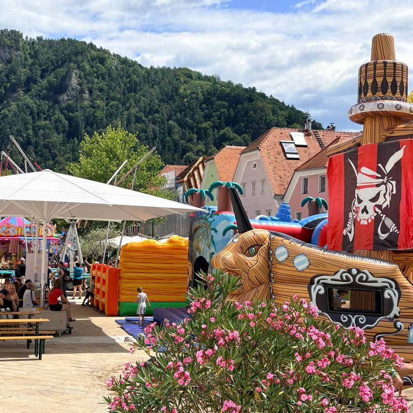 Eine farbenfrohe Außenszene mit aufblasbarem Piratenschiff und anderen Attraktionen, Menschen sitzen unter einem weißen Schirm, und ein Berg im Hintergrund.