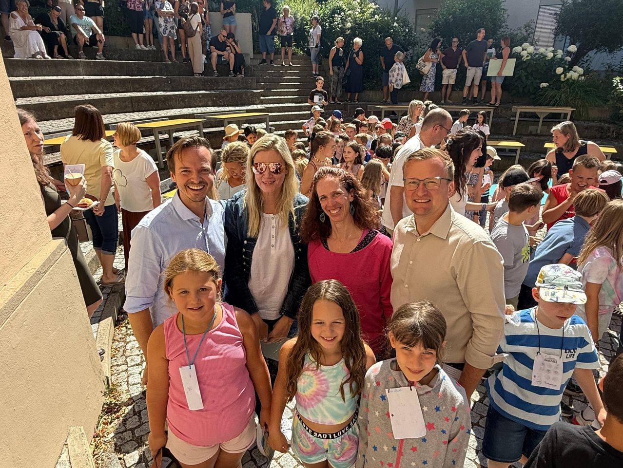 Eine Gruppe von Erwachsenen und Kindern, darunter zwei Frauen und zwei Männer, lächeln und posieren für ein Foto vor einer Menge. Sie stehen auf einer Kopfsteinpflasterstraße mit einer Kulisse aus Treppen und Grün.