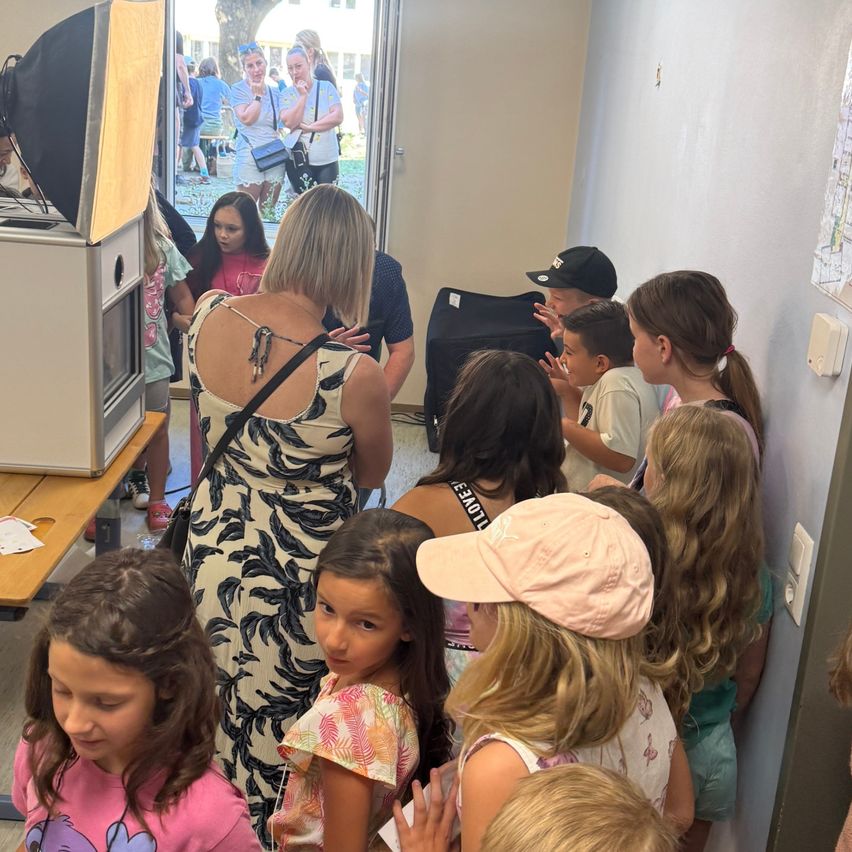 Eine Gruppe junger Mädchen und Jungen versammelt sich in einem Raum, wobei eine Frau in einem Blumenkleid in der Nähe eines Fotoautomaten steht. Ein Mann steht hinter dem Automaten und einige Leute stehen außerhalb des Raumes.