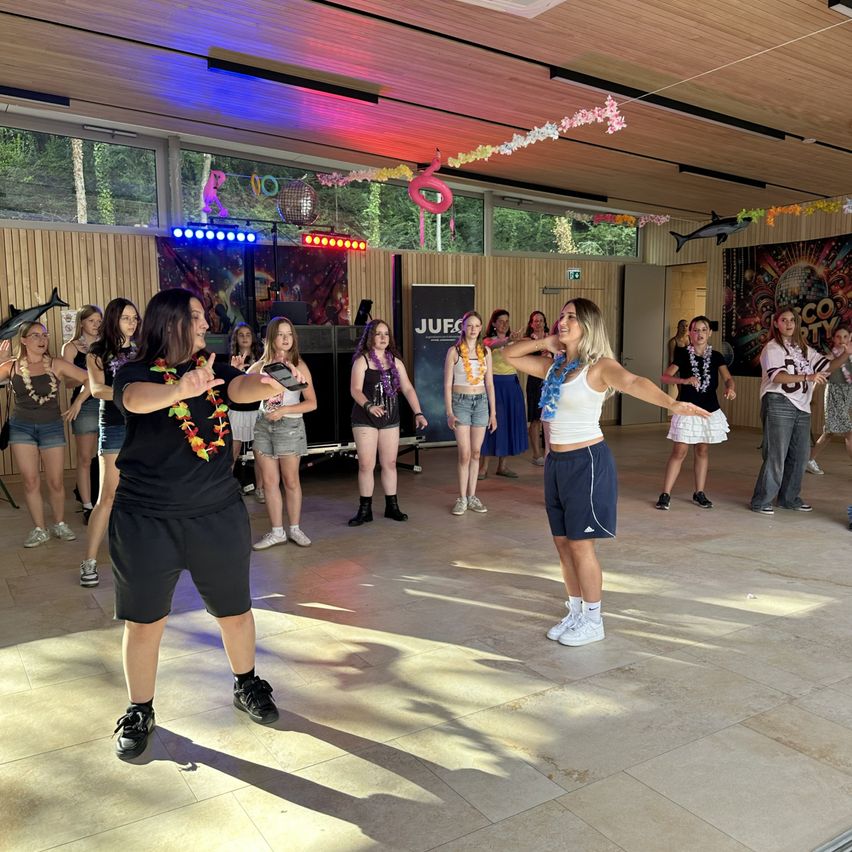 Eine Gruppe von Frauen in lässiger Kleidung tanzt in einem Raum mit Disco-Thema, mit bunten Lichtern und Dekorationen.