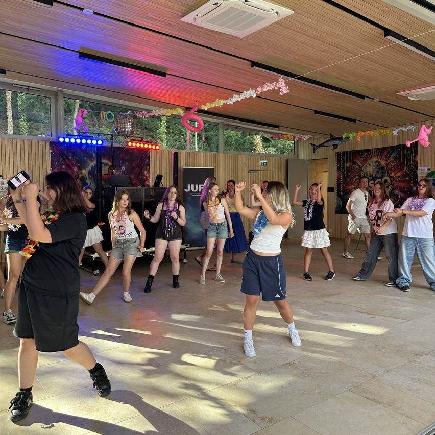 Eine Gruppe junger Frauen tanzt in einem Raum mit Disco-Lichtern und Dekorationen. Sie tragen Freizeitkleidung und einige haben Kränze um den Hals.