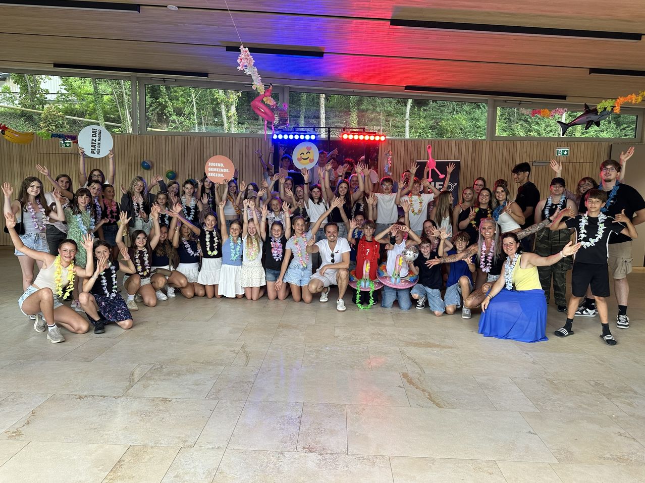 Eine Gruppe von Menschen, wahrscheinlich ein Team, posiert für ein Foto in einem Raum mit Disco-Licht-Setup, einige tragen Blumenkränze und halten Schilder hoch.