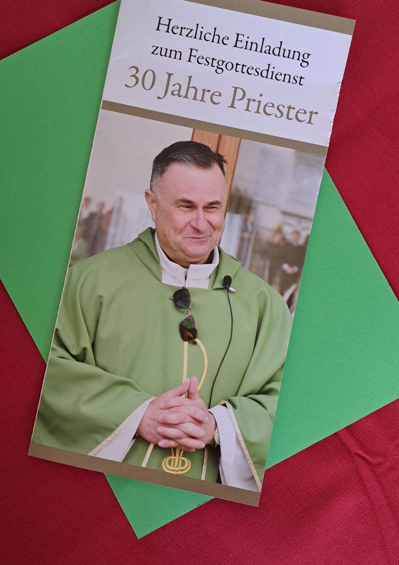 Ein Buch mit dem Titel '30 Jahre Priester', das einen lächelnden Priester mit einer um den Hals hängenden Sonnenbrille zeigt, liegt auf einem grünen und roten Hintergrund.