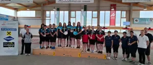 Eine Gruppe von Personen steht auf einem Podium, trägt Medaillen und scheint einen Sieg zu feiern. Sie befinden sich in einer Sporthalle, mit Bannern und Trophäen sichtbar.
