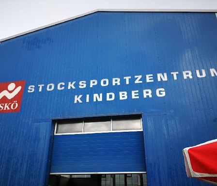 Ein blaues Gebäude mit weißem Text trägt die Aufschrift Stocksportzentrum Kindberg. Ein rotes und weißes Logo mit dem Wort Asko ist links. Ein roter und weißer Regenschirm befindet sich in der rechten Ecke.