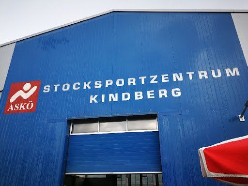 Ein blaues Gebäude mit weißem Text trägt die Aufschrift Stocksportzentrum Kindberg. Ein rotes und weißes Logo mit dem Wort Asko ist links. Ein roter und weißer Regenschirm befindet sich in der rechten Ecke.