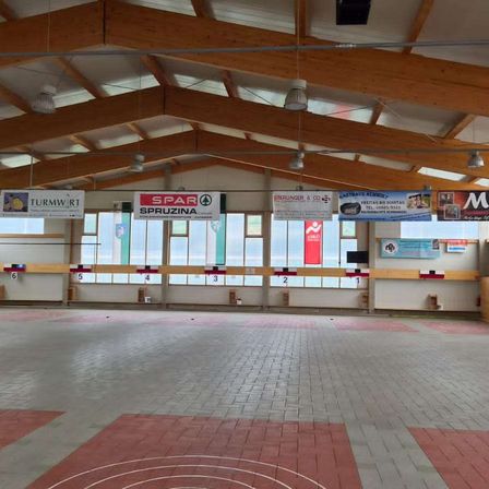 Ein Indoor-Bereich mit Ziegelboden, leerem Raum, hölzerner Dachkonstruktion, hängenden Lampen, Bannern mit Nummern und einem großen Glasfenster.